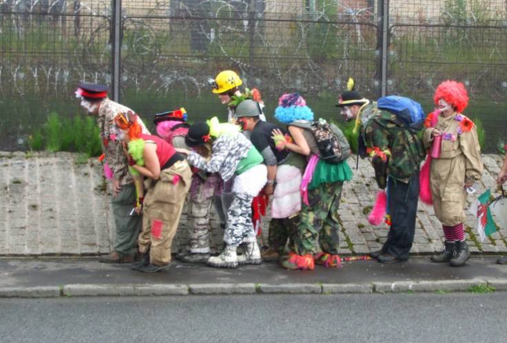 Bu görsel Gizli Palyaço Ordusu'nun (CIRCA) olduğu bir fotoğraf. Fotoğrafta beton ve tel örgülerden oluşan bir duvarın önünde sıraya girip eğilmiş 9 palyaço gözüküyor. Üsterine renkli parçalarla yamalanmış asker üniformaları giymişler. Pembe mavi sarı peruklar, asker şapkaları ve renkli baretler takmışlar. Palyaçolardan bir kısmı tel örgülerden içeri bakıyor, bir kısmı ise iler bakıyor. Eğilmeleri olduğunu anlamak ya da harekete geçmek için pozisyon aldıkları düşündürüyor.