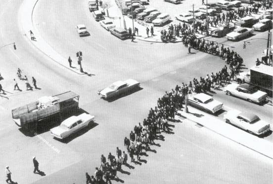 Bu görsel siya beyaz bir fotoğraf. Fotoğraf 50'lerin sonundaki Nashville eylemlerinden. Dört yolun bir ucundan diğer ucuna ikili sıraya girmiş ve yolu kapatmış bir grup insan gözükor. Bu insanların etrafında arabalar ve başka bir kaç insan yeralıyor.